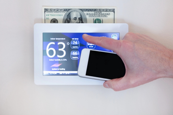man's hand holding a smart phone while adjusting home thermostat with dollar bill on top depicting rising cooling costs