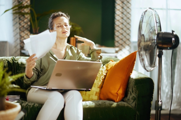 woman feeling hot and humid indoors while using paper as fan and indoor fan